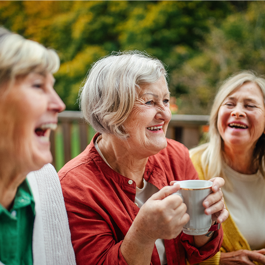 Three older women laughing outdoors with coffee mugs